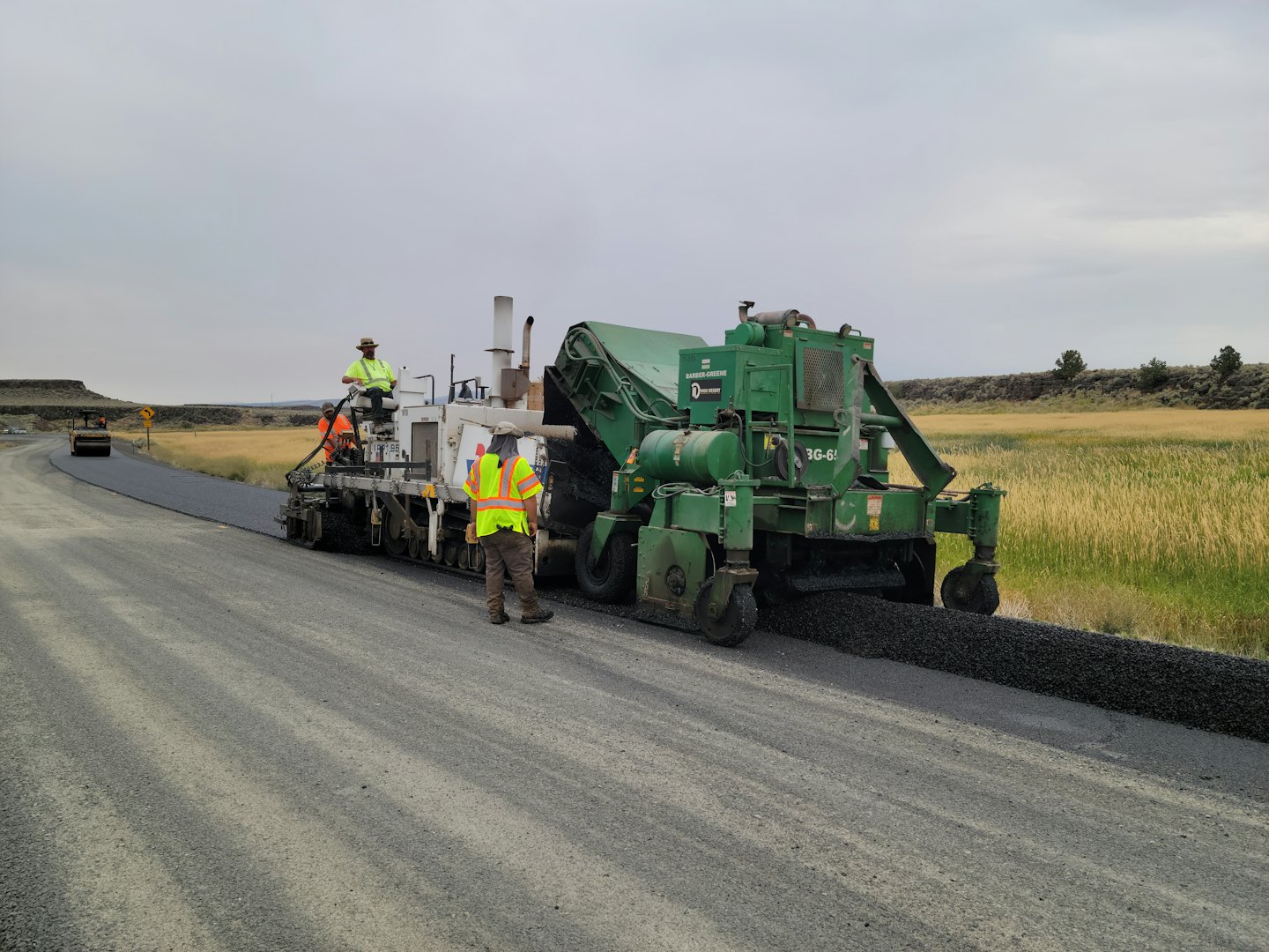 Construction workers pave a rural road with heavy machinery.